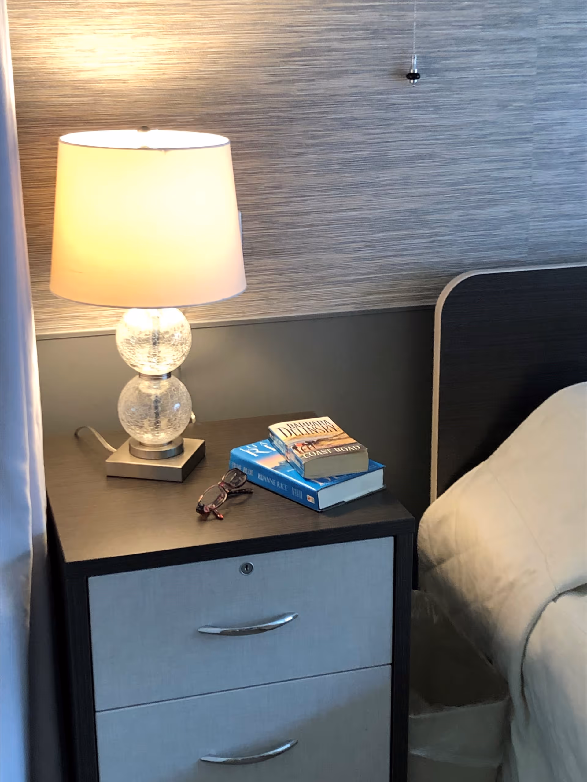 A bedside table with two drawers next to a bed. On the table is a lamp with a glass base, two books stacked on top of each other, and a pair of eyeglasses. The bed has a light-colored comforter and a dark headboard. The wall behind has a textured wallpaper with a pull cord hanging down.