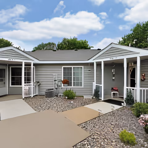 Exterior view of a single-story building with gray siding and a dark roof, featuring a small porch area with white railings and a ramp leading to the entrance. The surrounding area has landscaping with rocks and some greenery under a partly cloudy sky.