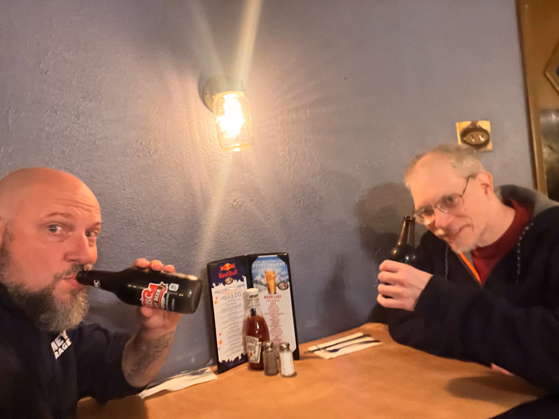 Two men sitting at a wooden table in a dimly lit room with a blue wall, each holding a dark bottle and drinking. On the table are condiments including ketchup, salt, and pepper shakers, along with a menu standing upright. A wall-mounted light fixture is above them.