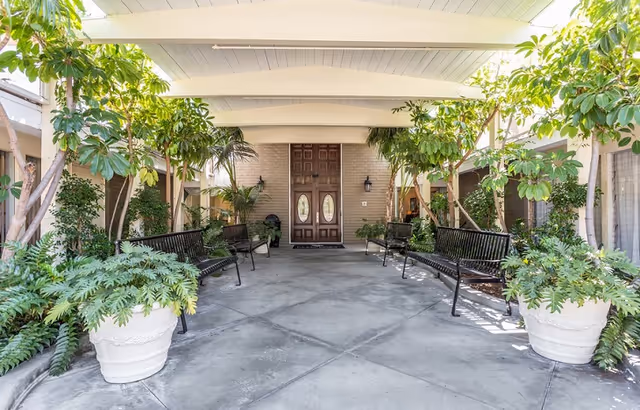 Covered outdoor seating area with black metal benches and large potted plants on either side, leading to a wooden double door entrance surrounded by greenery.