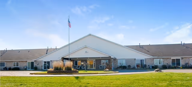 Front exterior view of a single-story senior living facility building with a central entrance, an American flag on a flagpole, and a well-maintained lawn under a blue sky with some clouds.