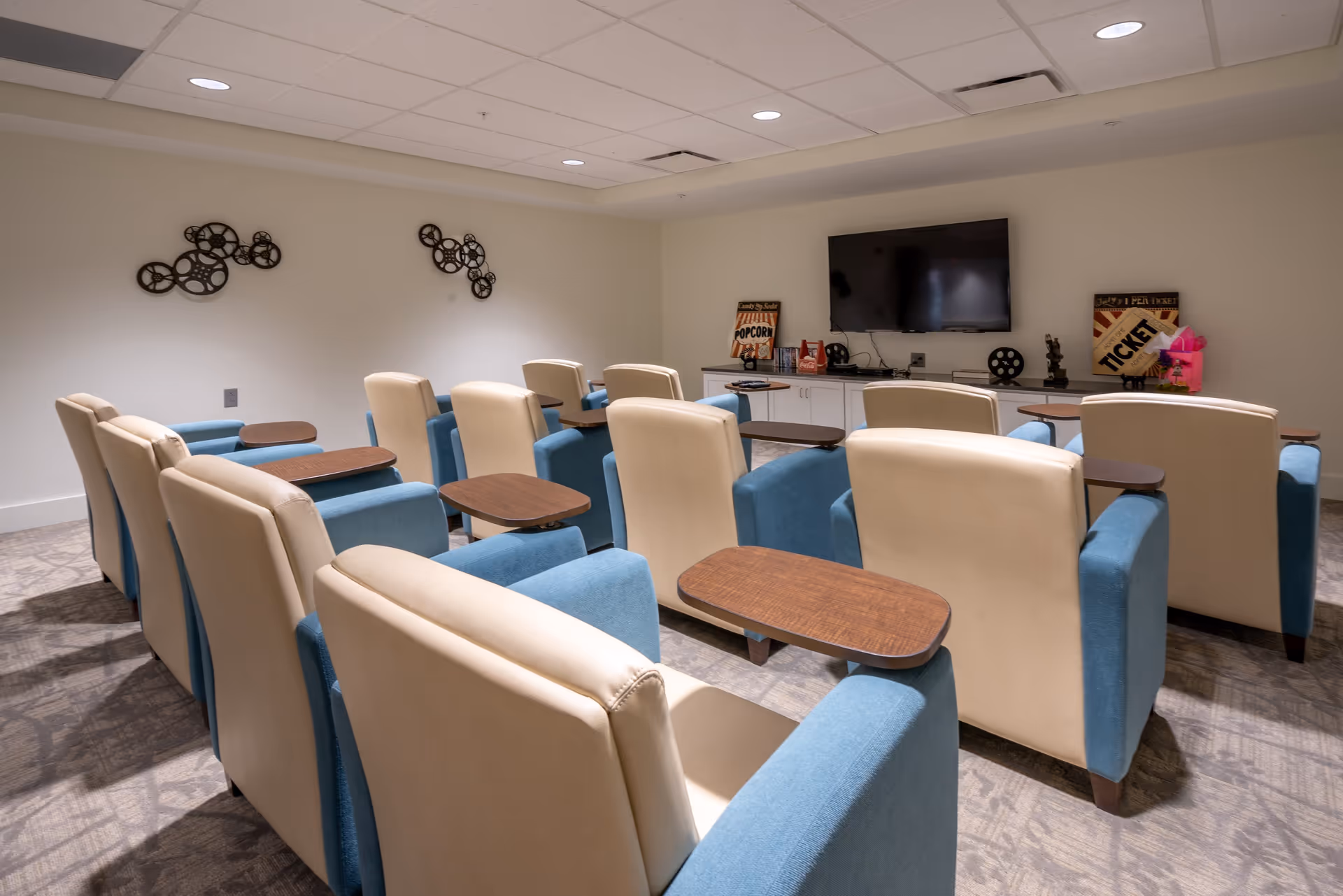 A small theater-style room with two rows of beige and blue armchairs, each equipped with a wooden tray table. At the front of the room is a flat-screen TV mounted on the wall above a white cabinet. The walls are decorated with movie-themed items including film reels and vintage-style popcorn and ticket signs.