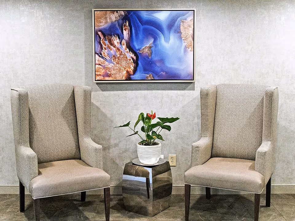Two beige upholstered armchairs with wooden legs are placed on either side of a small metallic hexagonal table holding a white pot with a green plant and red flowers. Above the chairs, a framed abstract artwork with blue and brown tones hangs on a light gray wall.