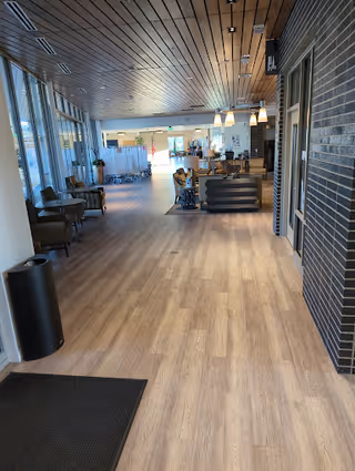 A spacious hallway in a senior living facility with light wood flooring and a wooden ceiling. On the left side, there are large windows letting in natural light and several chairs arranged along the wall. On the right side, there is a dark brick wall. In the distance, there are tables and chairs, some partitions, and hanging light fixtures.