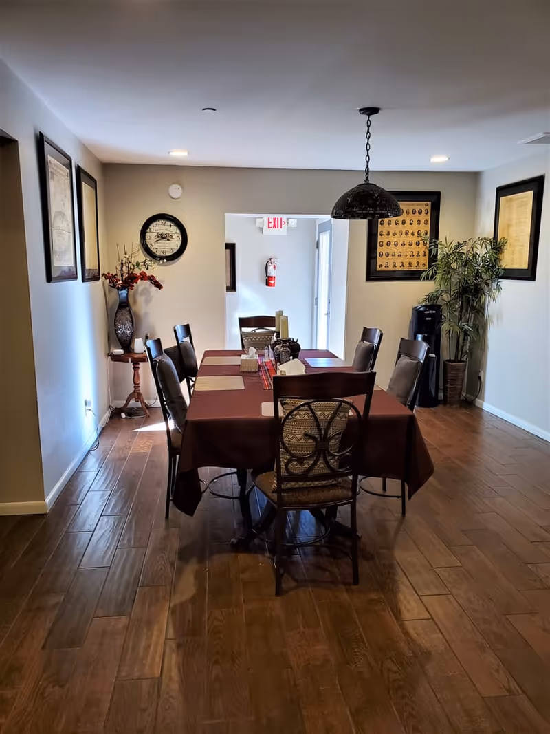 A dining room with a rectangular table covered with a dark red tablecloth and surrounded by eight chairs. The room has wooden flooring, framed pictures and a clock on the walls, a vase with flowers on a small side table, a hanging light fixture above the table, and a potted plant near the corner. An exit sign and fire extinguisher are visible in the background.