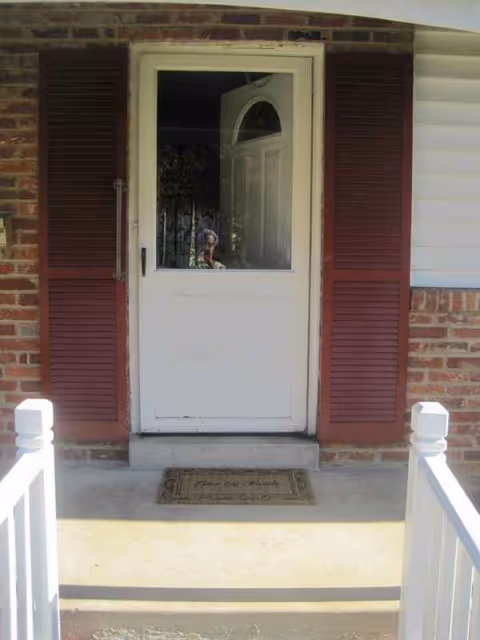 White front door with maroon shutters on brick exterior, entry mat, and white railing.