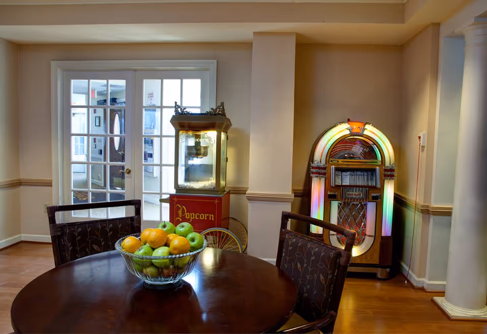 Interior room with a round wooden table and two chairs. On the table is a glass bowl filled with green apples and oranges. In the background, there is a vintage popcorn machine and a colorful jukebox against the wall. French doors with glass panes lead to another room.