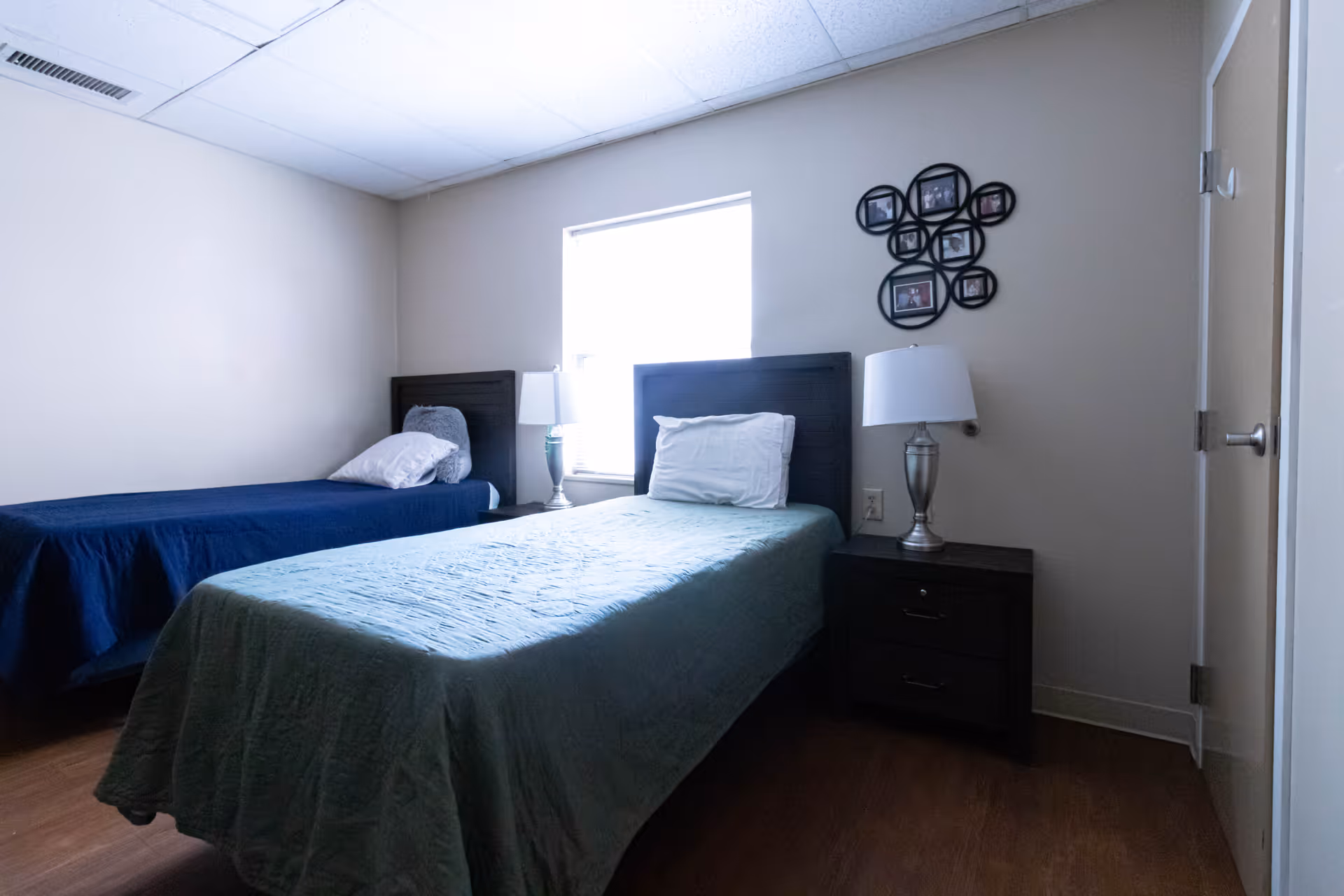A simple bedroom with two single beds, one with a green bedspread and the other with a blue bedspread. Each bed has a pillow and a headboard. There is a window between the beds letting in natural light. A nightstand with a lamp is positioned between the beds, and a decorative photo frame hangs on the wall above the nightstand. The room has light-colored walls and a wooden floor.