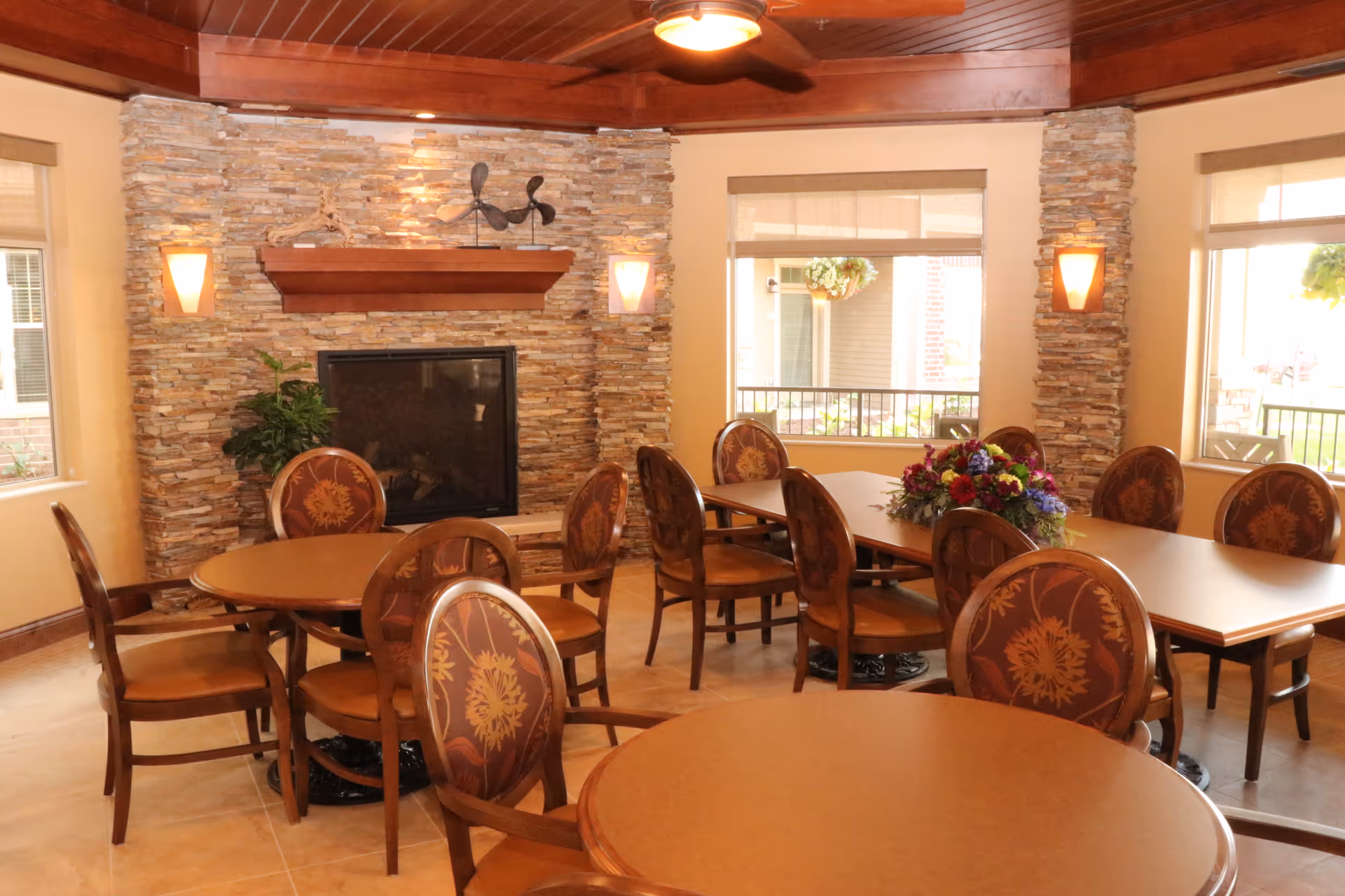 A cozy dining area with round and rectangular wooden tables surrounded by cushioned chairs with floral upholstery. The room features a stone fireplace with a wooden mantle, wall sconces, large windows with beige blinds, and a wooden ceiling with a ceiling fan. A floral centerpiece decorates one of the tables.