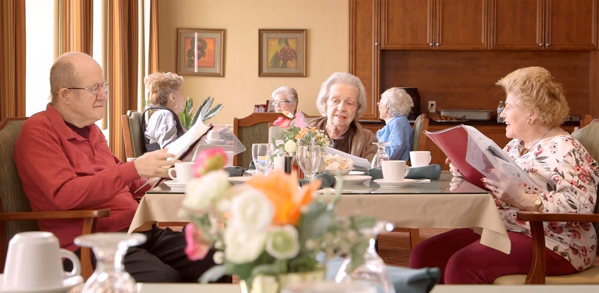 A group of elderly people sitting around a dining table in a well-lit room, reading menus and engaging in conversation. The table is set with cups, glasses, and a floral centerpiece. The background shows wooden cabinets and framed artwork on the walls.