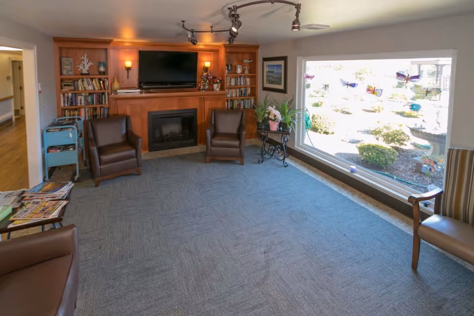 A cozy living room area with a large window overlooking a garden. The room features a fireplace with a TV mounted above it, flanked by built-in bookshelves filled with books and decorative items. There are three brown leather armchairs arranged around the fireplace, a small table with magazines, and a plant stand with potted plants near the window. The floor is carpeted and the room is softly lit with wall sconces and track lighting.