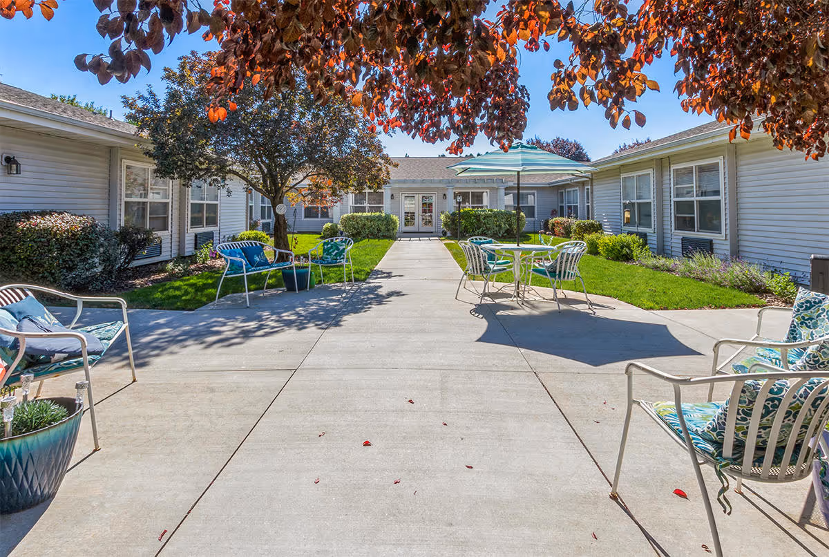 Outdoor courtyard area at Avista Senior Living Garden City with a concrete walkway, green grass, trees with red leaves, and several metal chairs and tables with cushions and an umbrella providing shade.