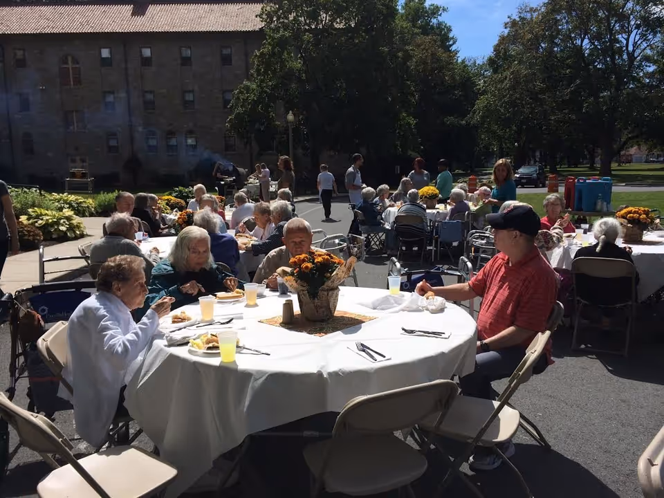 Seniors seated at round tables outdoors eating under sunny skies with a large stone building and trees in the background.
