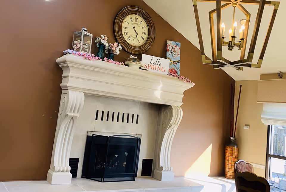 A cozy living room corner featuring a large white ornate fireplace with a black screen in front. On the mantel are decorative items including a round clock, flower vases, a lantern, and a sign that reads 'hello SPRING'. The wall behind the fireplace is painted brown, and a large window with a beige shade lets in natural light. A decorative vase with tall sticks stands in the corner, and a brass chandelier with exposed bulbs hangs from the ceiling.