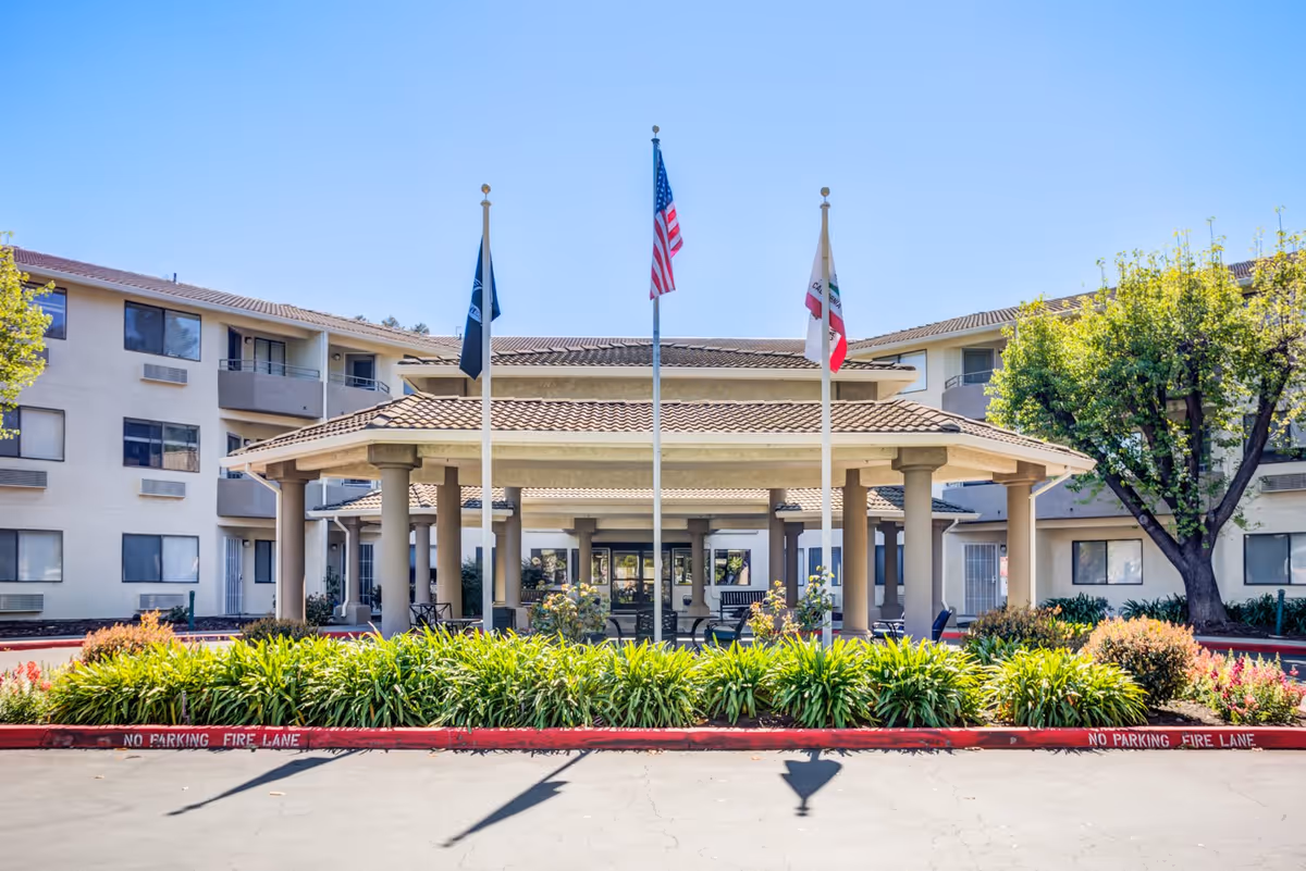 Front exterior view of Truewood by Merrill, Modesto senior living facility with a covered entrance supported by columns, three flagpoles displaying flags, landscaped greenery, and a clear blue sky.