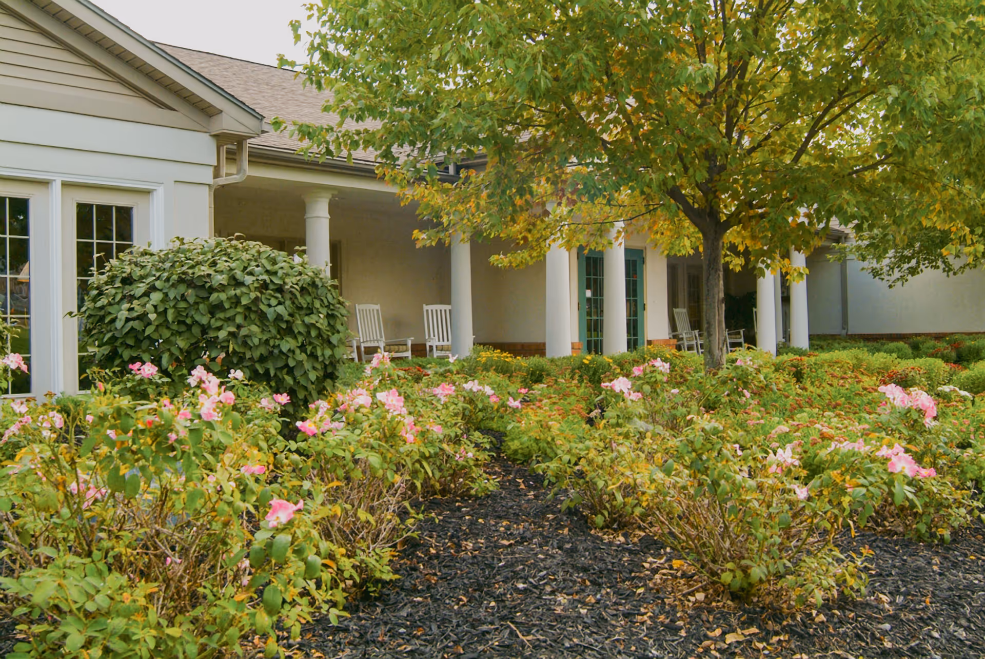 Outdoor garden area with blooming pink flowers and green shrubs in front of a building with white columns and a porch featuring white rocking chairs. A tree with green and yellow leaves is also visible.