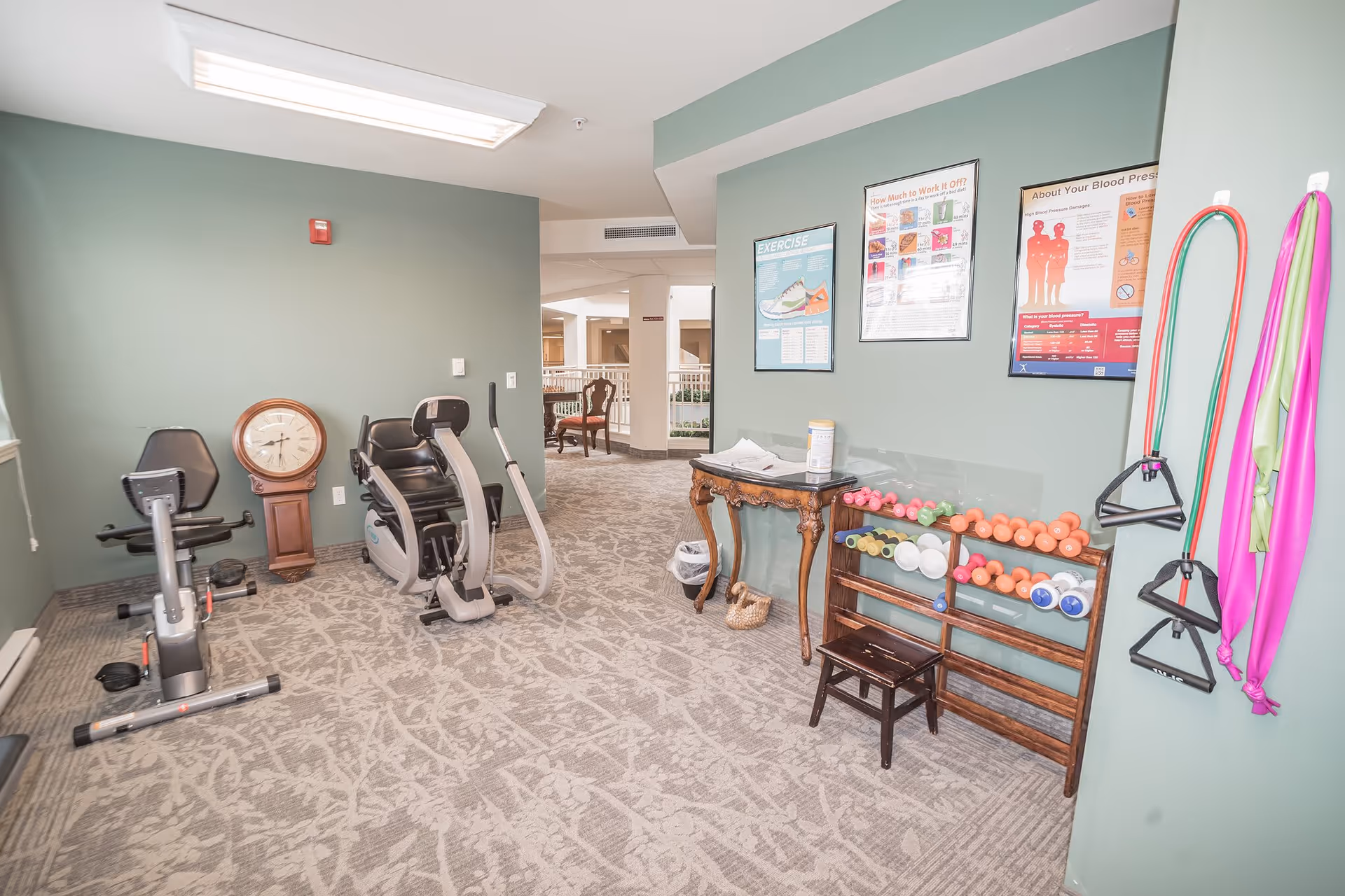 A small exercise room with light green walls and patterned carpet. The room contains two exercise bikes, a wooden clock, a rack with various dumbbells, resistance bands hanging on the wall, and a small wooden table with cleaning supplies. Three informational posters about exercise and health are displayed on the wall.
