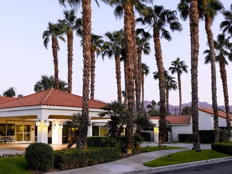 Exterior view of a senior living facility with white buildings featuring red tile roofs, surrounded by tall palm trees and well-maintained green landscaping at dusk.