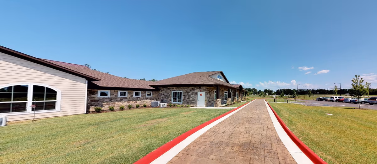 Exterior view of a single-story building with stone and siding walls, a brown shingled roof, and several windows. A paved walkway with red curbs runs alongside the building, leading to a parking lot with several cars. The sky is clear and blue.