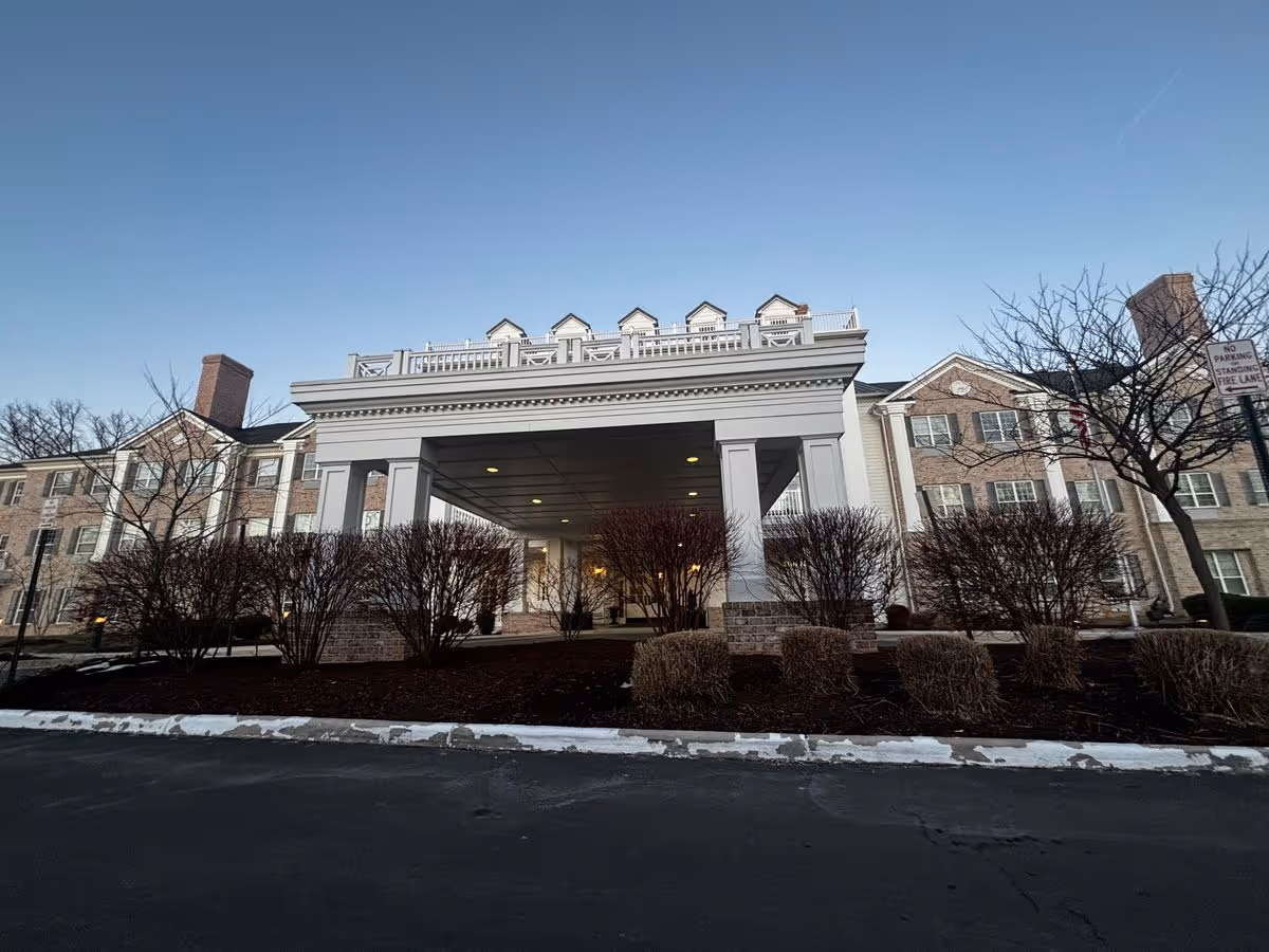 Front entrance of a multi-story brick senior living building with a covered porte-cochere and trimmed leafless shrubs under a clear sky.
