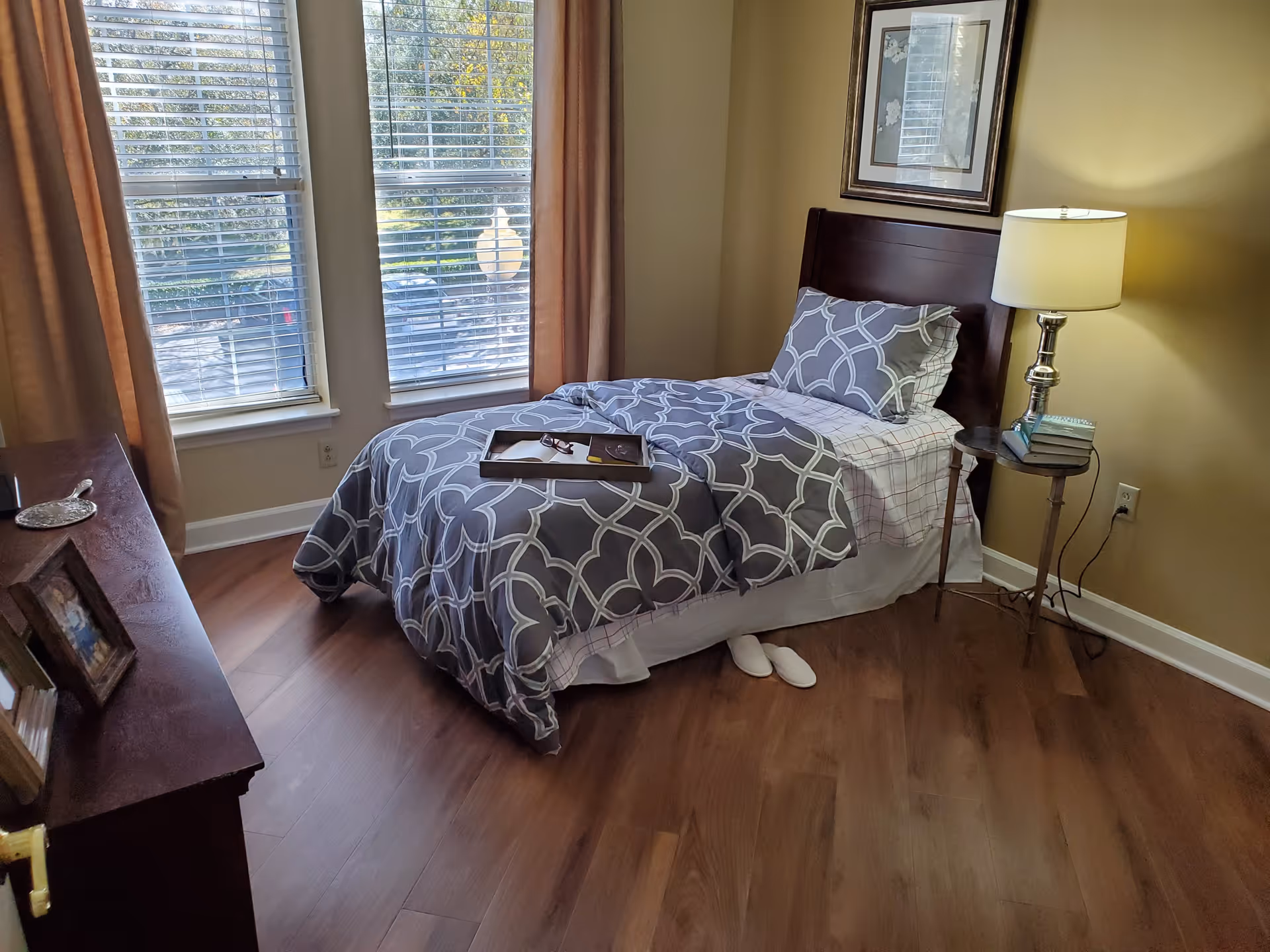 A cozy bedroom with a single bed covered in a gray and white patterned comforter and matching pillow. A wooden headboard is visible, along with a small round side table holding a lamp and some books. There are two large windows with blinds and beige curtains, and a wooden dresser with framed photos on the left side. The floor is wooden, and a pair of white slippers is placed under the bed.