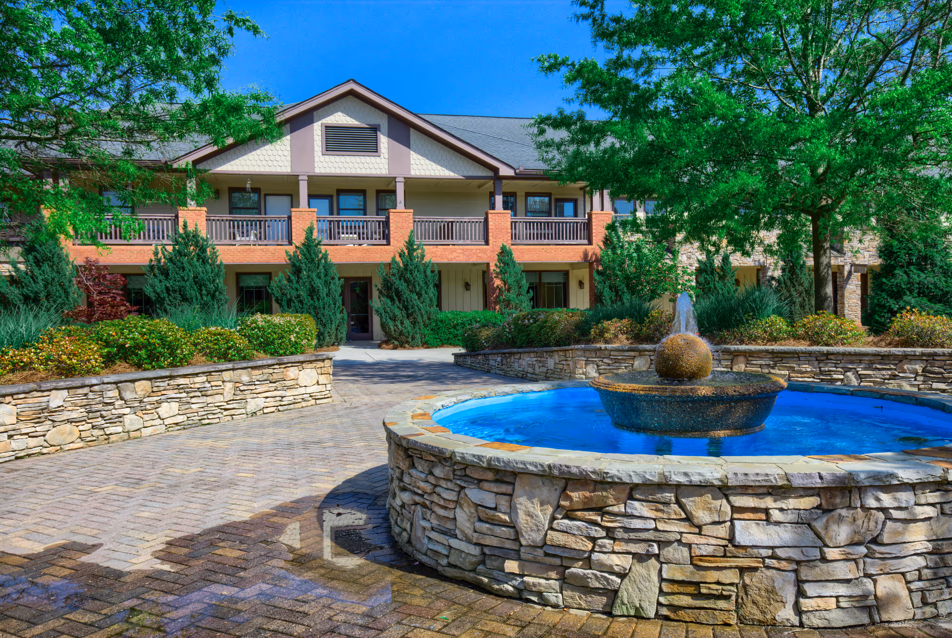 Outdoor view of TerraBella Sugarloaf facility showing a stone fountain with water in the foreground, surrounded by a paved walkway and landscaped greenery. The building has two stories with balconies and is set against a clear blue sky.