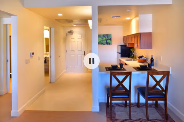 Interior view of a senior living facility apartment showing an entryway with a white door, a small kitchen area with wooden cabinets, a countertop with two place settings, and two wooden chairs. The walls are painted light beige, and there is a small piece of artwork on the kitchen wall.