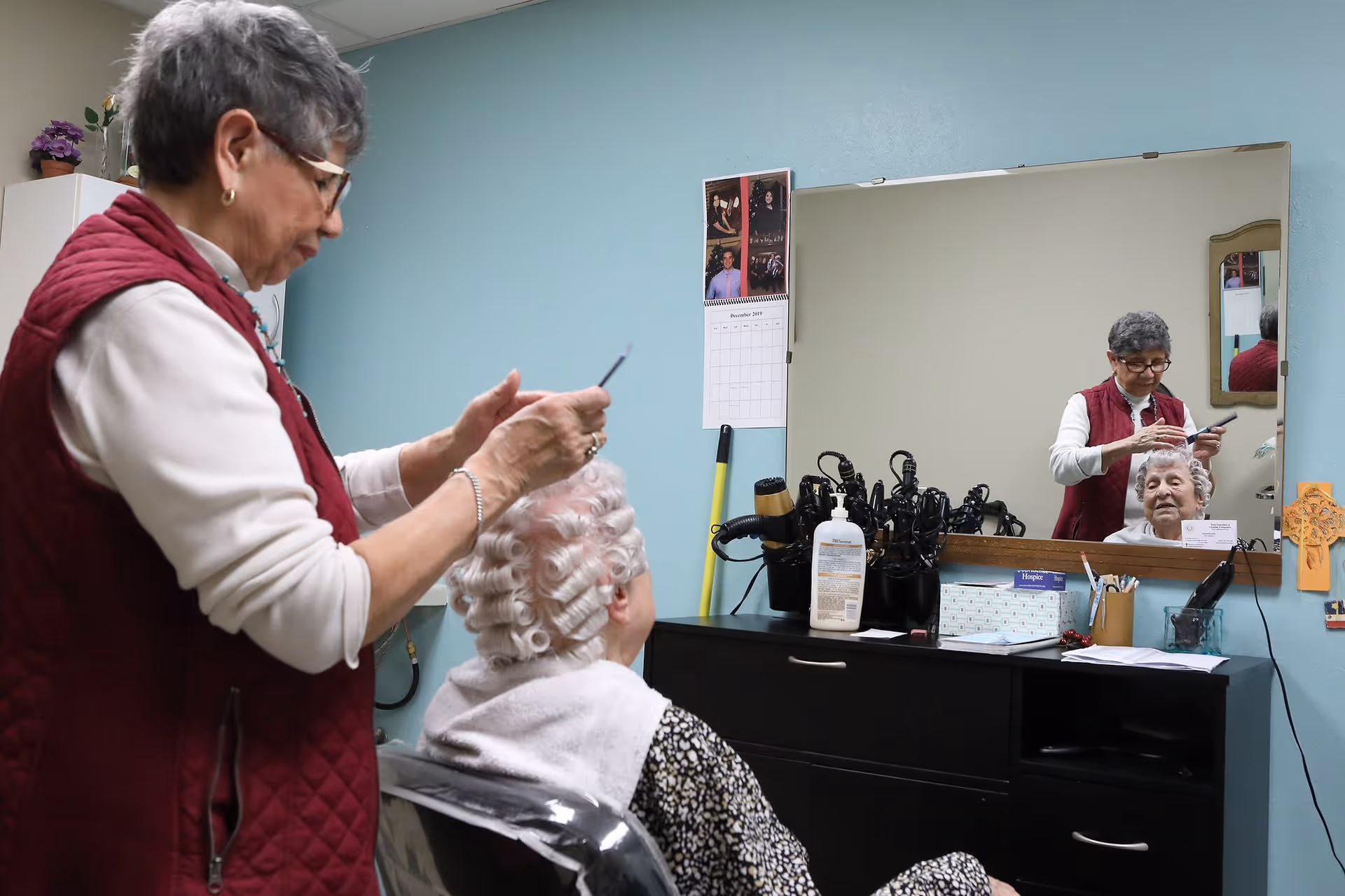 An elderly woman is seated in a salon chair with curlers in her hair while another woman, possibly a stylist, is standing next to her holding a comb. They are in a room with a large mirror, a cabinet with hair styling tools, and a calendar on the wall.