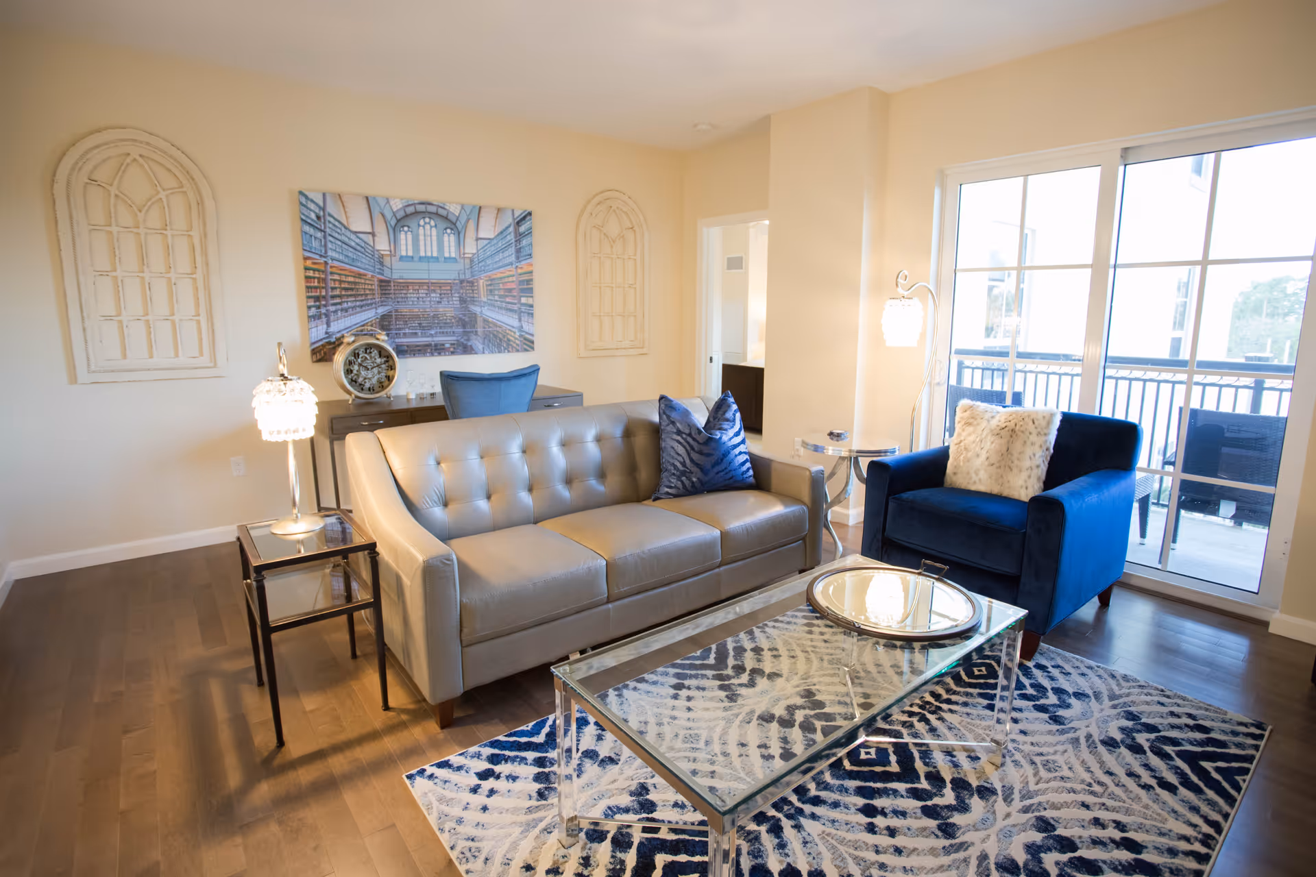 A bright living room with a gray tufted sofa adorned with a blue pillow, a blue armchair with a white furry pillow, a glass coffee table on a patterned blue and white rug, two side tables with lamps, a large window with sliding glass doors leading to a balcony, and wall decor including a large picture of a library and two decorative window frames.