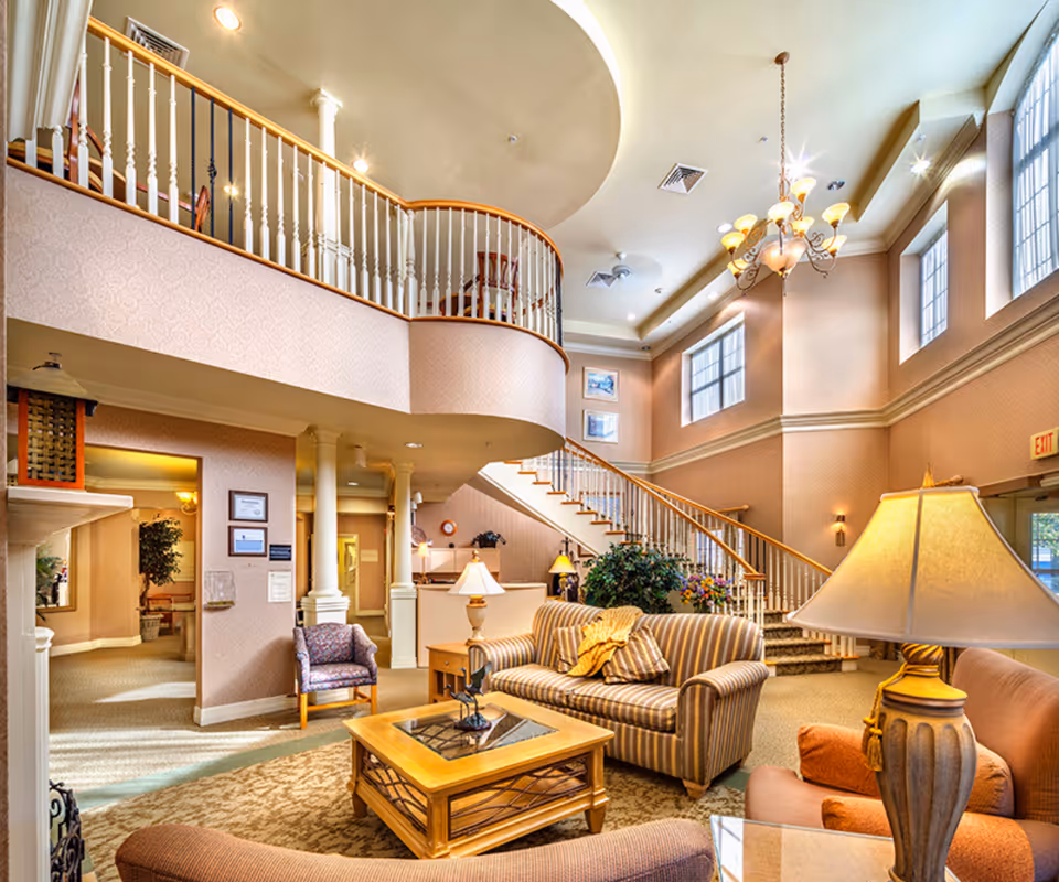A spacious and well-lit living room area in a senior living facility with high ceilings, large windows, and a chandelier. The room features comfortable seating including a striped sofa, armchairs, and a wooden coffee table. A staircase with white railings leads to an upper level balcony. The walls are decorated with framed pictures and the space includes lamps and plants for a cozy atmosphere.