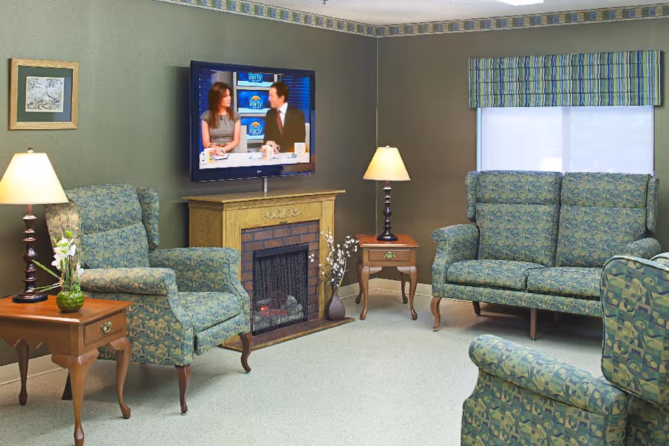 Comfortable living room with patterned armchairs and a loveseat, a TV mounted above a fireplace, lamps and side tables.