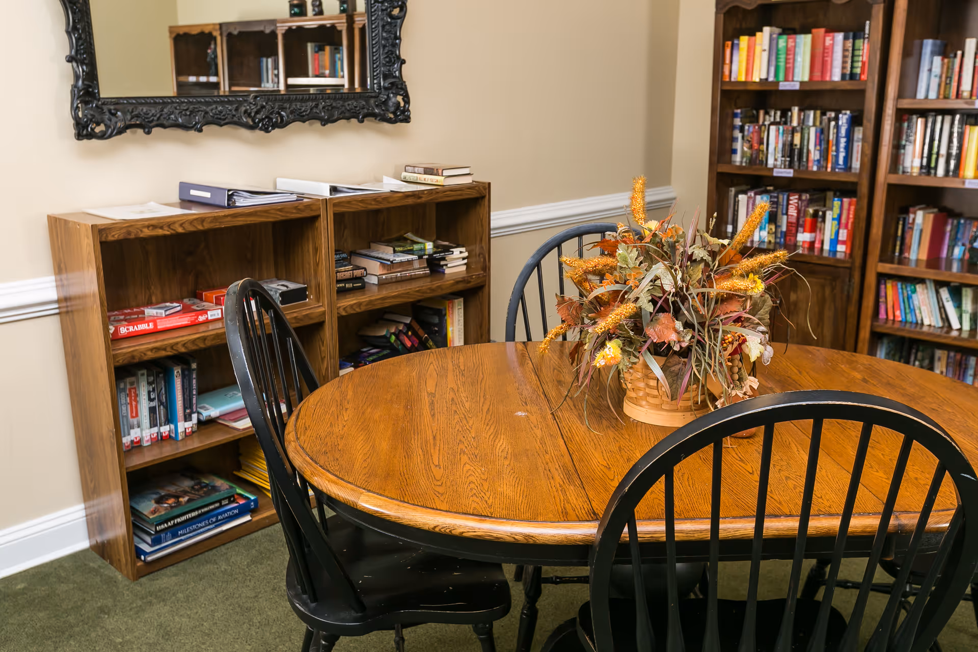A cozy interior room with a round wooden table surrounded by black wooden chairs. On the table is a decorative basket with autumn-themed artificial flowers. Behind the table are wooden bookshelves filled with books and board games. A large ornate mirror hangs on the wall above the bookshelves.