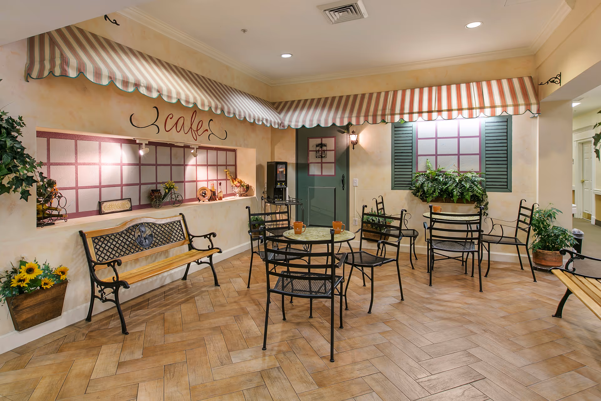Indoor cafe area with striped awnings, a green door, decorative window shutters, and several black metal tables and chairs. There are two wooden benches with metal armrests, potted plants, and a coffee machine on a small table. The walls are painted in warm tones with decorative accents and the word 'cafe' written above a serving window.