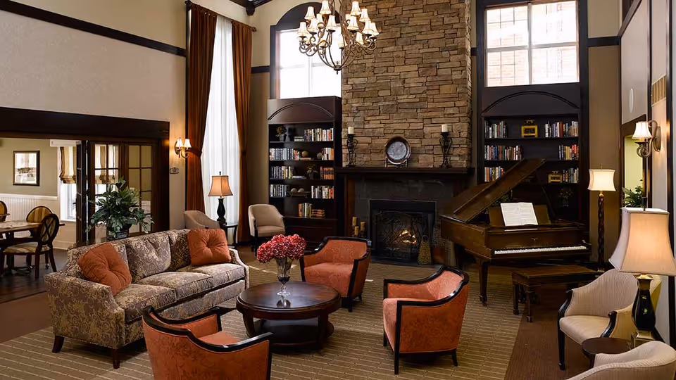 A cozy living room with a patterned sofa, four orange armchairs, a round wooden coffee table with a vase of flowers, a grand piano with sheet music, two tall bookshelves flanking a stone fireplace with lit candles, and large windows with brown curtains letting in natural light.