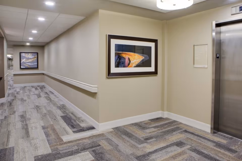 Interior hallway of a senior living facility with beige walls, patterned carpet flooring, handrails along the walls, two framed abstract paintings, and a stainless steel elevator door on the right.