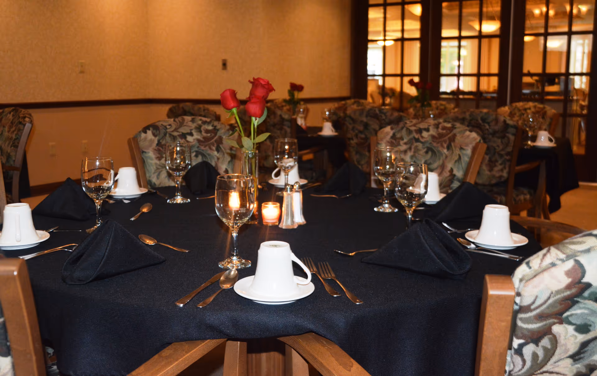 A dining table set for six with black tablecloth, black folded napkins, white cups on saucers, silverware, wine glasses, a small candle, and a vase with red roses in a senior living facility dining room with floral upholstered chairs and glass-paneled doors in the background.