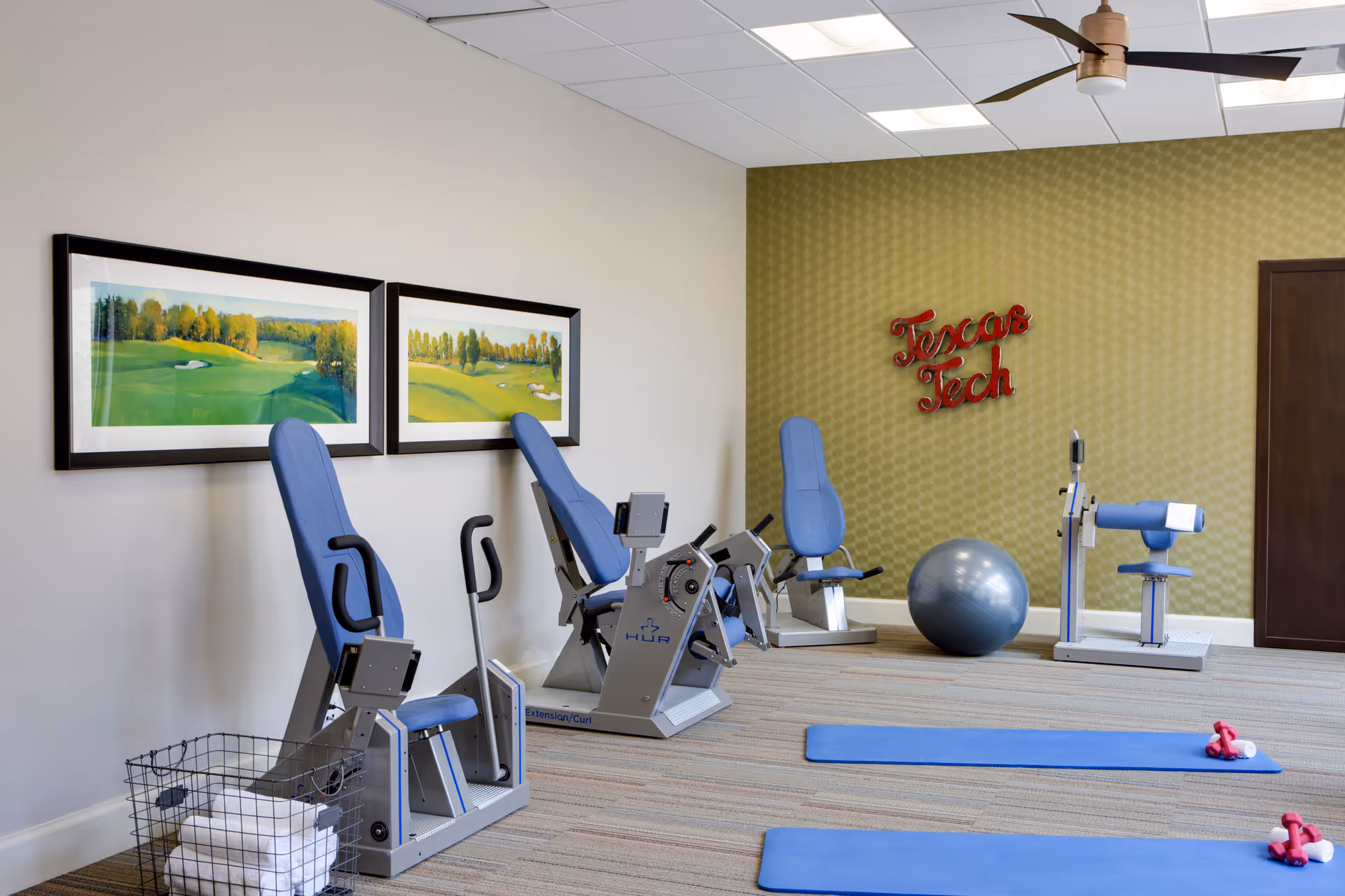 A small fitness room with exercise equipment including three blue padded workout machines, two blue exercise mats with pink dumbbells and white towels on them, a large silver exercise ball, and a wire basket with folded white towels. The walls are decorated with two framed landscape paintings and a red neon sign that reads 'Texas Tech'.