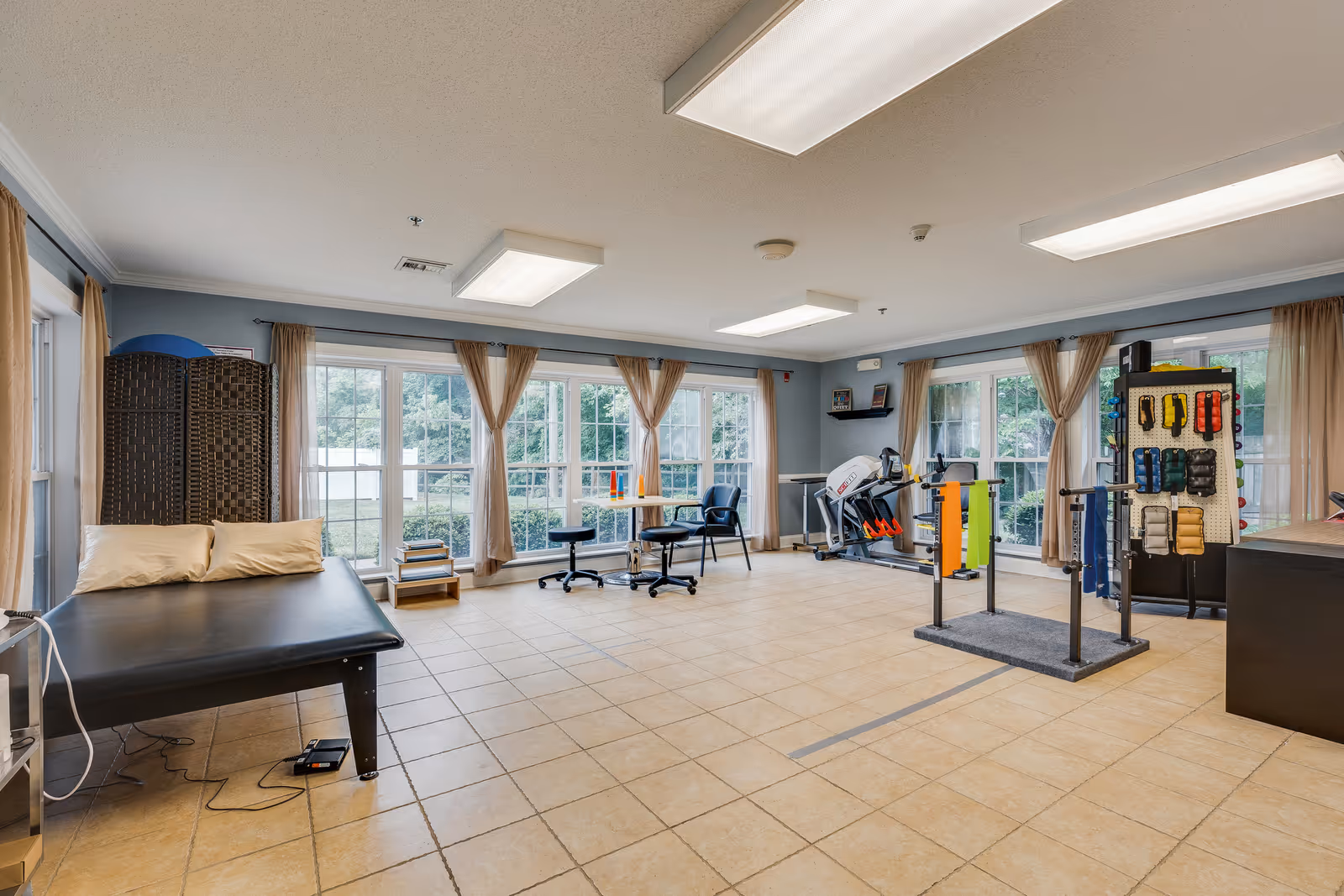 A spacious room with large windows covered by beige curtains, allowing natural light to fill the space. The room features a black therapy or massage table with two pillows, a small table with two chairs, exercise equipment including a stationary bike, parallel bars for walking support, and a rack holding various exercise bands and weights. The floor is tiled, and the walls are painted light blue.