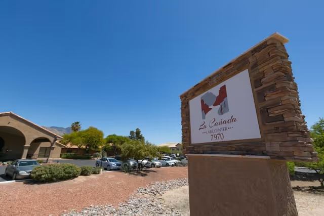 Stone entrance sign for La Cañada Care Center with the facility buildings and parked cars visible under a clear blue sky.