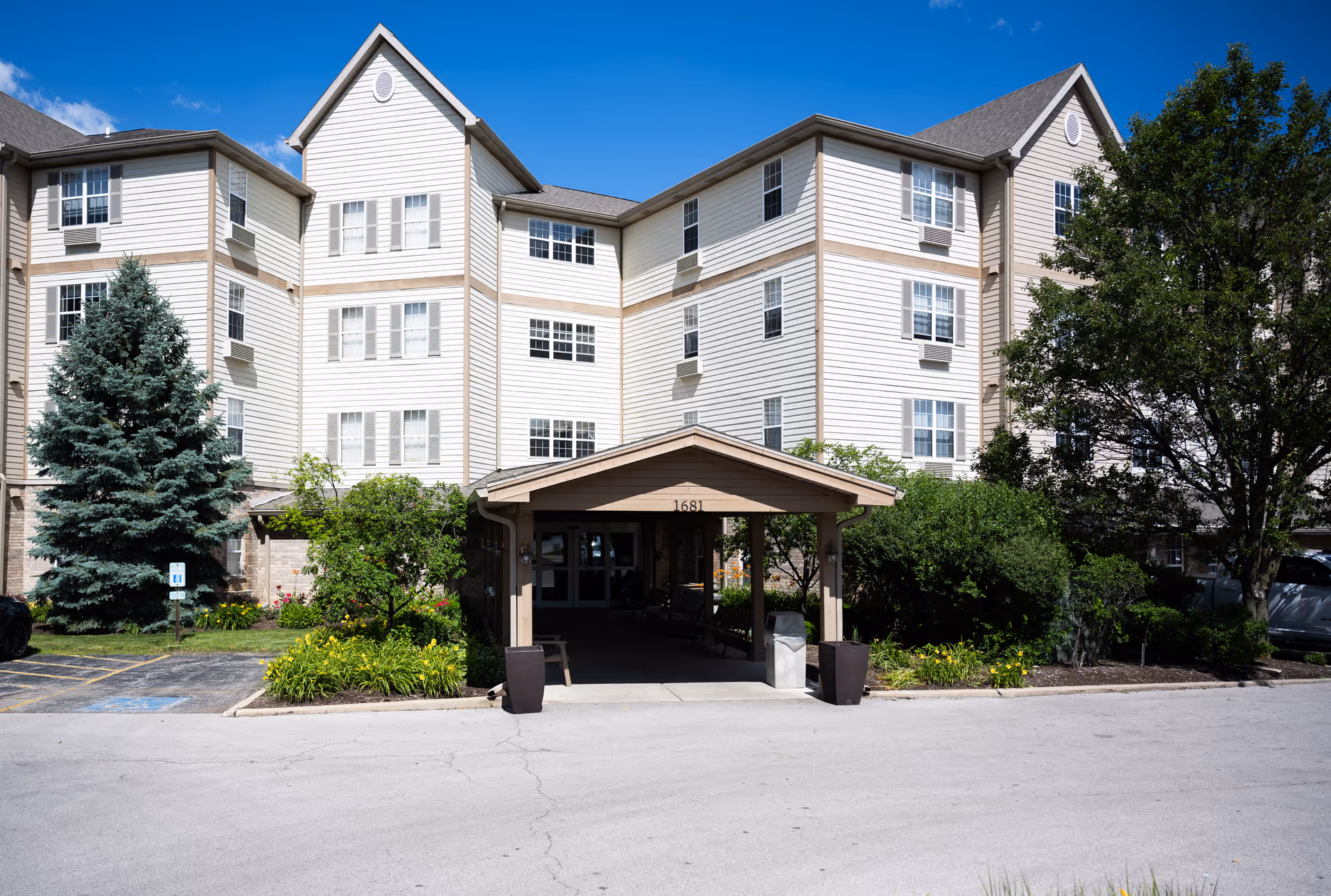 Front exterior of a multi-story beige senior living building with a covered entrance, landscaped beds, and surrounding trees.