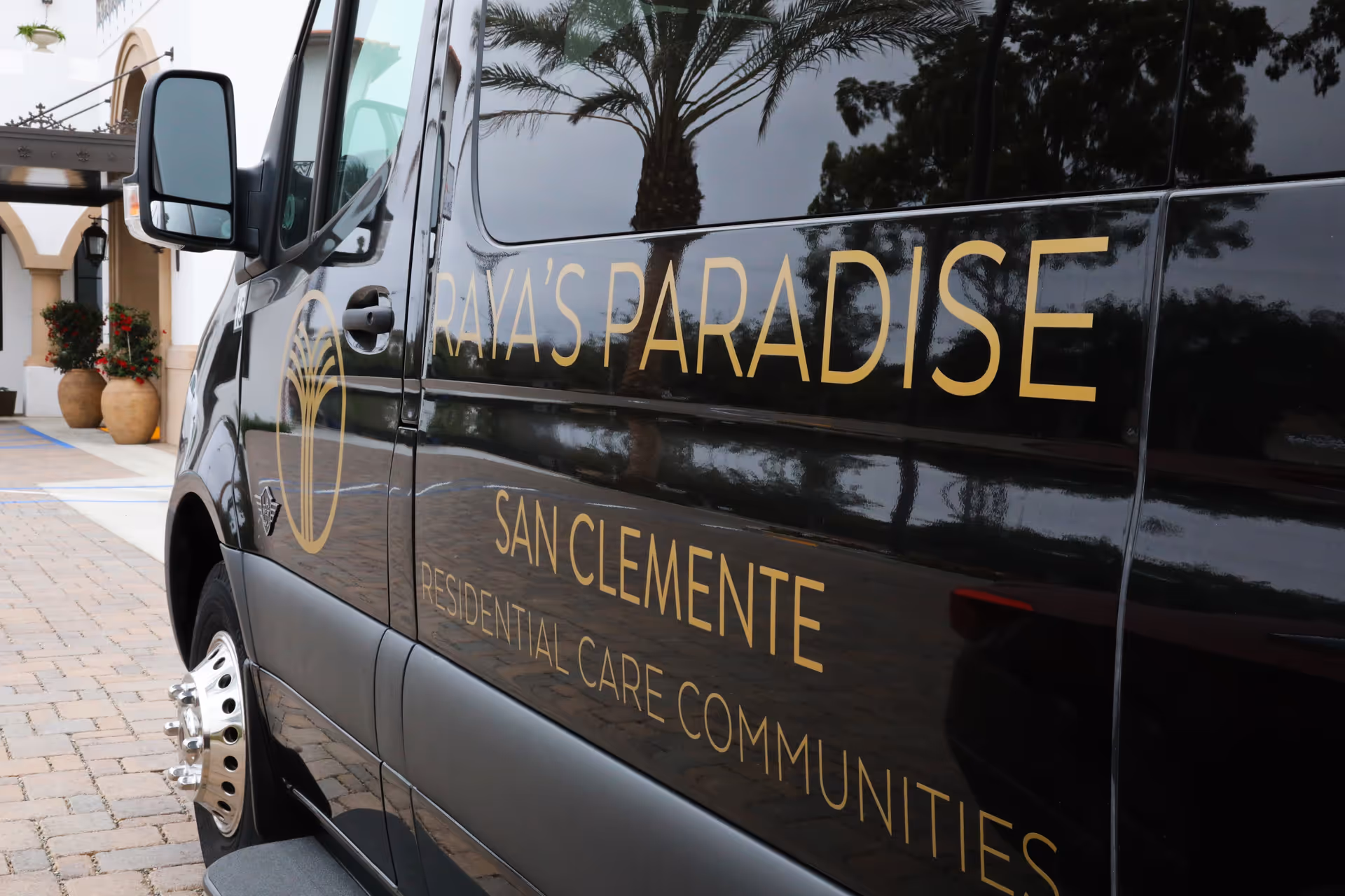 Close-up view of a black vehicle with gold lettering that reads 'Raya's Paradise San Clemente Residential Care Communities' parked on a brick driveway near a building entrance with large potted plants and arches.