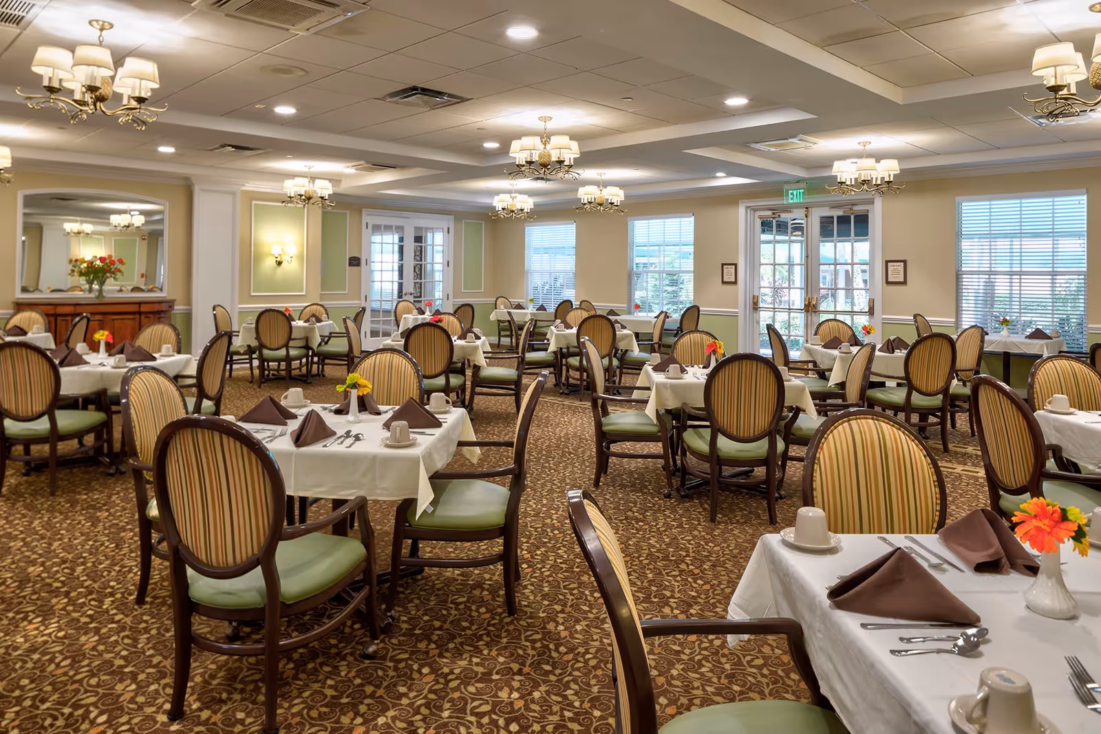 A spacious dining room with multiple tables covered in white tablecloths, each set with brown folded napkins, cups, and silverware. The chairs have wooden frames with striped upholstery and green cushions. The room features patterned carpet, chandeliers, wall sconces, large windows with blinds, and double glass doors leading outside.