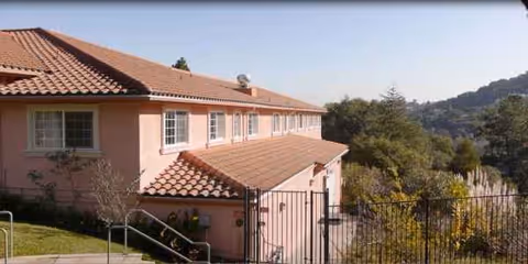 Pink stucco senior living building with terracotta roof on hillside, railing and windows.