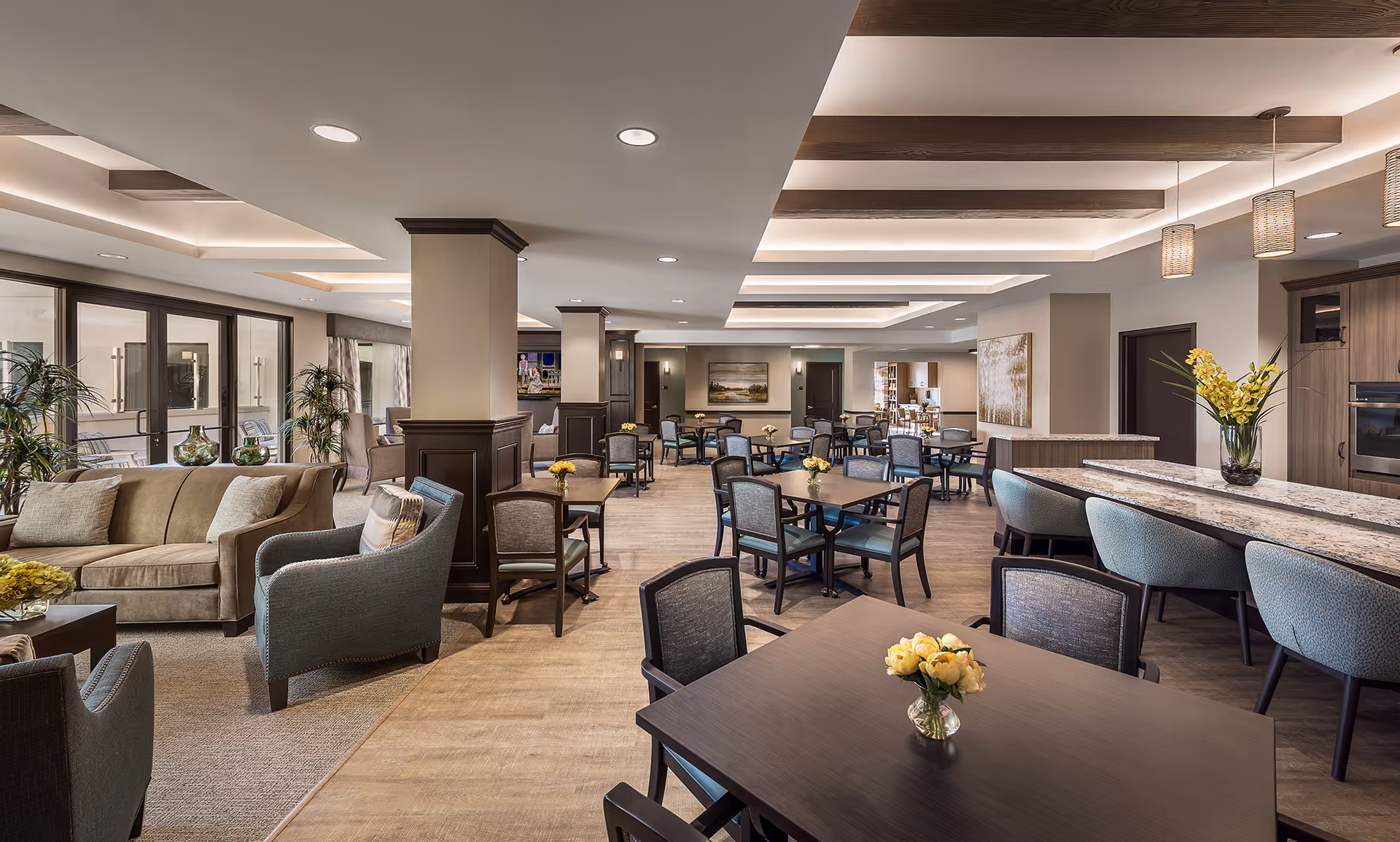 A spacious and well-lit common area in a senior living facility featuring multiple tables with chairs, a comfortable seating area with sofas and armchairs, decorative plants, and a countertop with bar stools. The ceiling has recessed lighting and wooden beams, and the room has large windows allowing natural light to enter.
