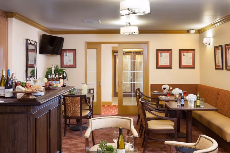 A cozy dining area in The Cottages of Carmel featuring a dark wooden bar with bottles of wine and beer, a small table with chairs, and a larger seating area with a long cushioned bench and multiple chairs. The room has warm lighting, framed artwork on the walls, and sliding glass doors leading to another room.