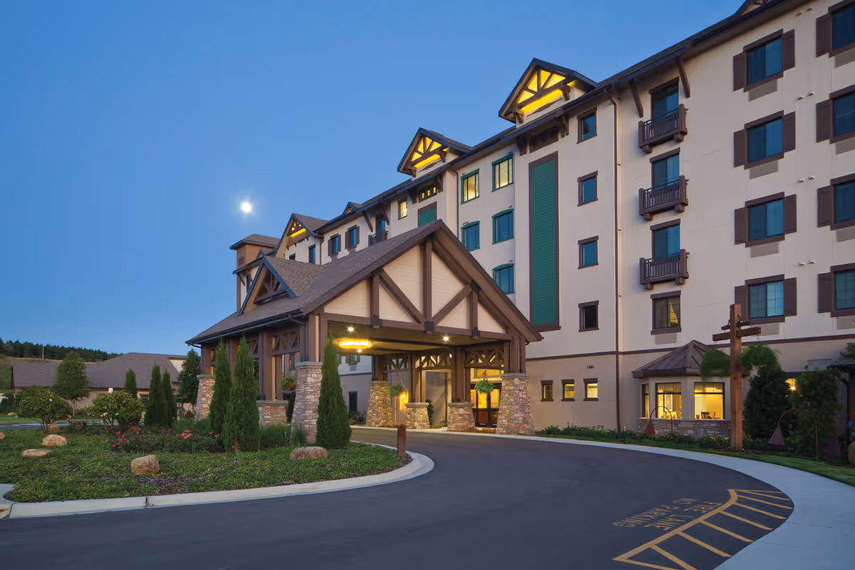 Front exterior of a multi-story senior living building with a covered porte-cochère entrance lit at dusk.