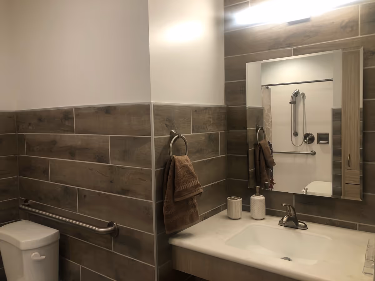 A bathroom with wood-look tile walls, a white toilet with a metal grab bar, a sink with a marble countertop, a mirror above the sink, a towel ring with a brown towel, and a shower with a handheld showerhead and grab bar visible in the mirror.
