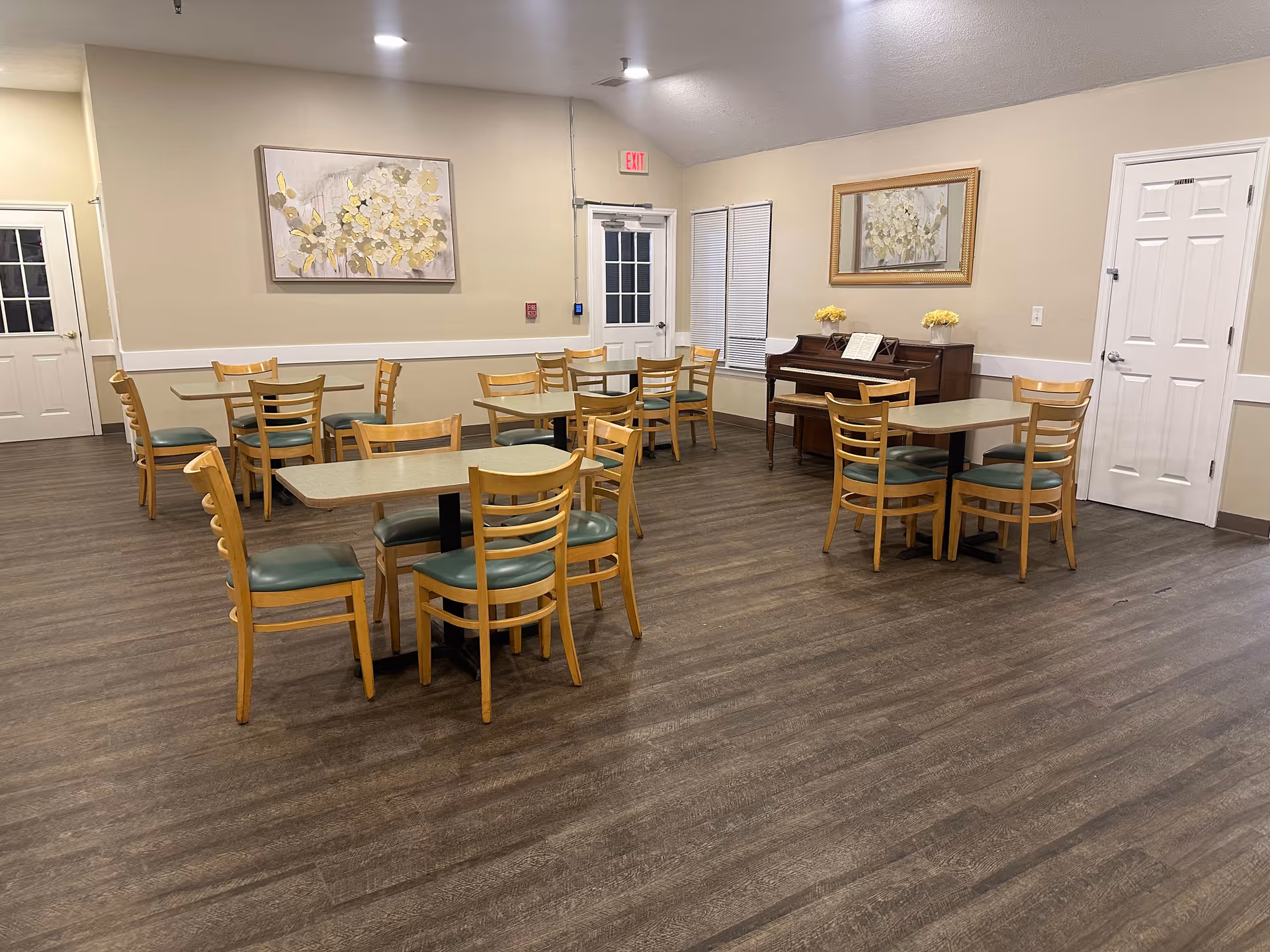 A dining area with several wooden tables and chairs arranged on a wood-patterned floor. The walls are beige with white trim, decorated with floral paintings. There is a piano against one wall with a music book and yellow flowers on top. Multiple doors and windows are visible, and the room is well-lit with ceiling lights.