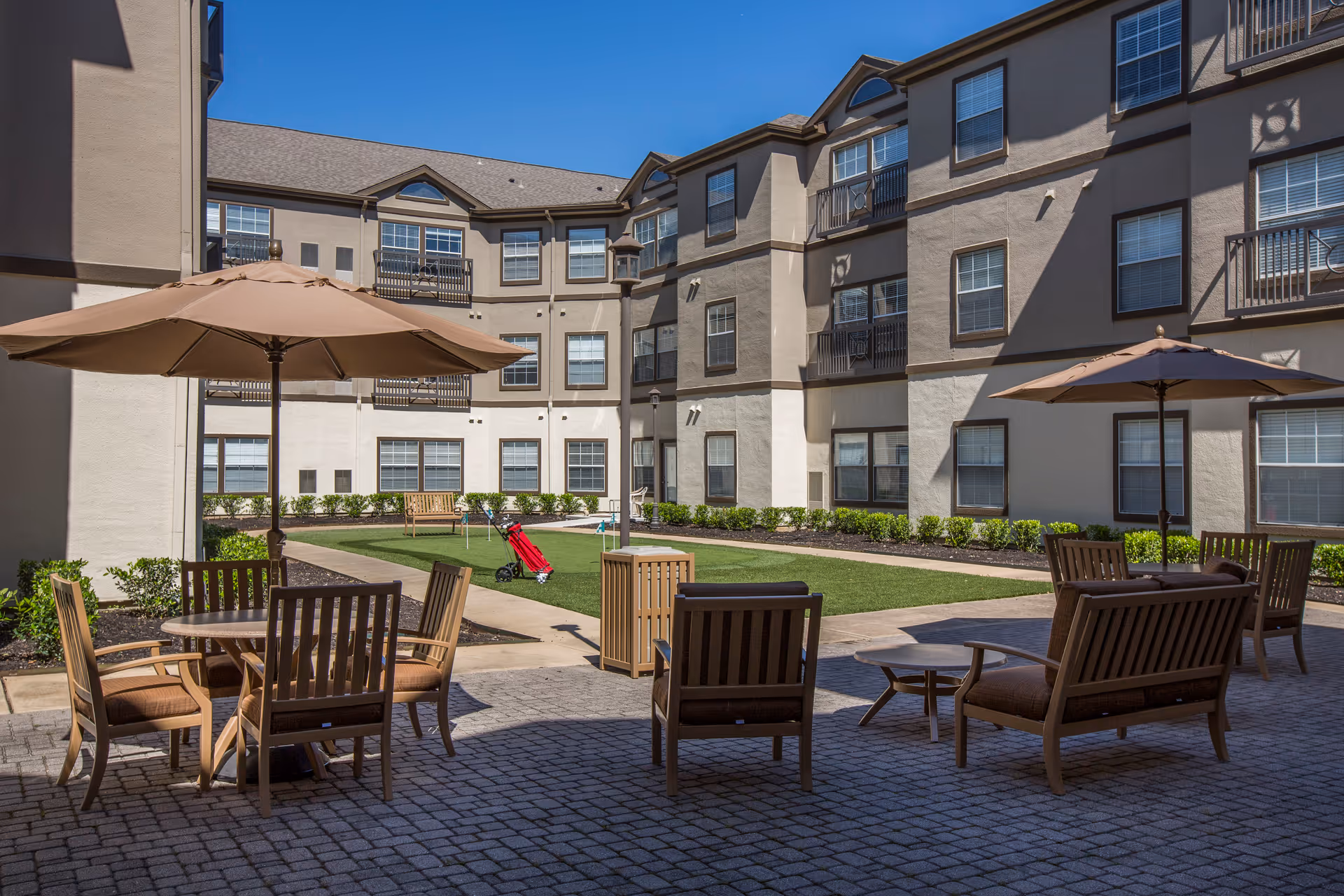 Outdoor courtyard area of a senior living facility with patio tables, chairs, umbrellas, and a small putting green surrounded by a three-story building under a clear blue sky.