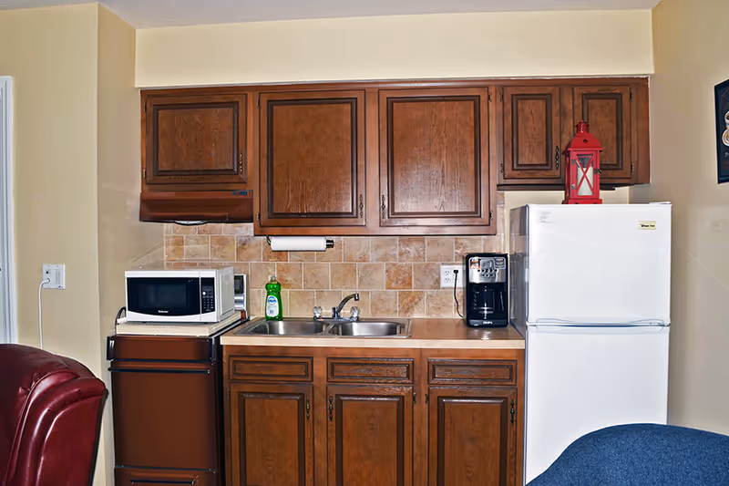 A small kitchen area with wooden cabinets, a double sink, a white microwave on a brown stove, a bottle of dish soap, a black coffee maker, and a white refrigerator with a red lantern on top. The backsplash is tiled in beige tones.
